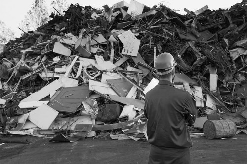 A man wearing a safety helmet and dark work clothing stands with his back to the camera, observing a large pile of mixed construction and industrial waste materials. The waste includes broken panels, metal scraps, plastic and cardboard debris, and other discarded building materials, all piled haphazardly in an outdoor setting on a paved surface. The collection appears unmanaged, with various textures and finishes visible, such as smooth plastic, corrugated cardboard, and rough metal edges. The environment suggests an industrial or waste management site, possibly near a commercial or construction area in Kilburn, where rubbish removal services by Commercial Waste Kilburn might be required. The lighting is diffuse, with no direct sunlight, emphasizing the chaotic nature of the waste pile and highlighting the contrast between the clean safety gear of the worker and the disorderly heap of refuse in the background.