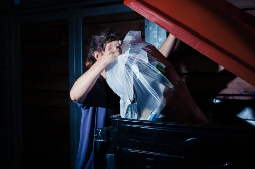 A person with short dark hair wearing a dark T-shirt is seen disposing of a large transparent plastic bag filled with mixed waste into a black skip or dumpster located in an outdoor or semi-covered area. The background features a wooden wall with horizontal panels and a metal framework painted in blue. The skip is positioned on a surface that appears to be paved or concrete, and the lid of the skip is open, revealing the interior. The image suggests an active waste disposal process, typical of commercial rubbish removal services, with focus on the handling of waste in an organised manner. The setting hints at a commercial or industrial environment associated with waste management or rubbish collection in Kilburn or the surrounding area, aligning with services offered by Commercial Waste Kilburn.