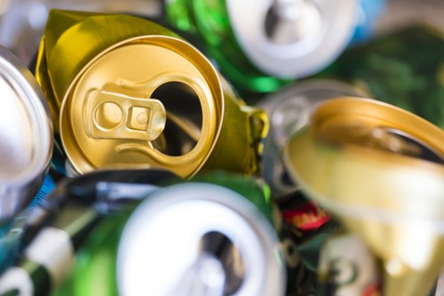 A close-up view of several crushed aluminium cans in various shades of metallic silver, gold, and green. The cans are tightly packed together, with some partially open and showing their pull-tab mechanisms, on a surface that appears to be part of a collection of waste materials. The textured surfaces of the cans reveal their typical aluminum finish, with some showing minor dents from crushing. The background is blurred, suggesting an indoor environment or a collection area used for rubbish collection and disposal. The composition emphasizes the metallic materials and the condition of used cans, aligning with a waste management context such as commercial waste removal services provided by Commercial Waste Kilburn, focusing on the disposal of beverage packaging.