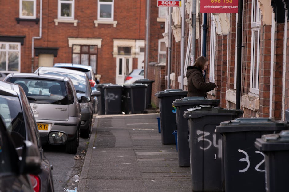 A black wheeled rubbish bin positioned on a sidewalk next to a curb at night, with its lid open and filled with various waste items including cardboard boxes and paper. The bin has a label with the te
