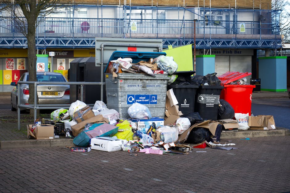 A large pile of mixed waste and rubbish is accumulated around a central metal recycling bin on a paved urban roadside. The waste includes various cardboard boxes, plastic bags, paper items, and packag